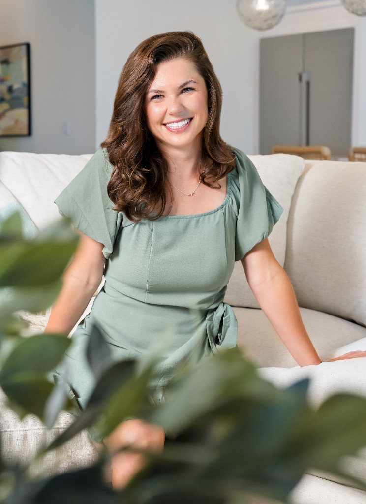 Professional portrait of a woman in a sage green dress seated on a sofa with soft natural light and greenery