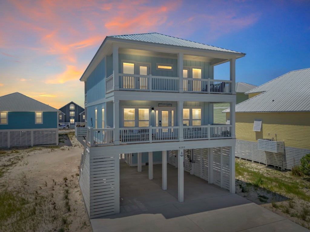 Light blue coastal beach house on stilts at sunset with warm interior lighting and a vibrant orange sky